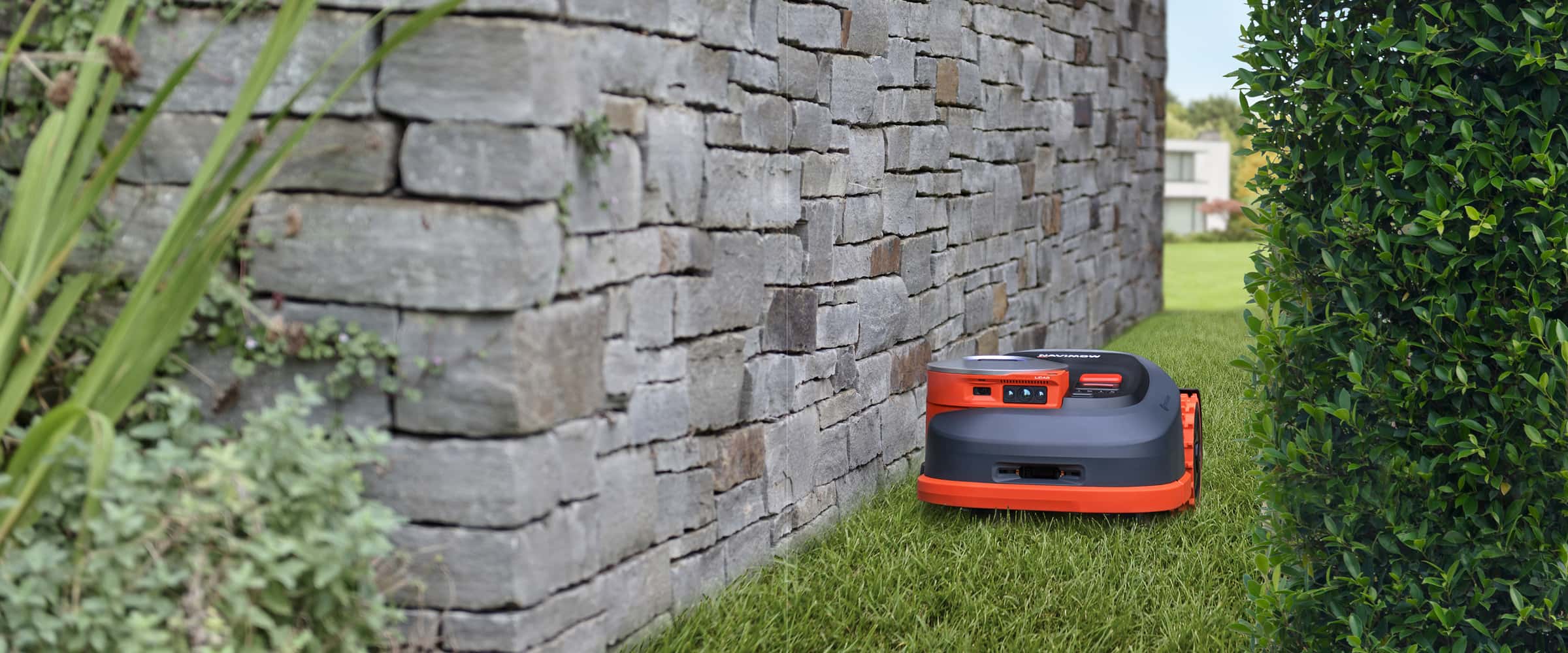 Navimow i110N robot lawn mower mows grass near stone wall.